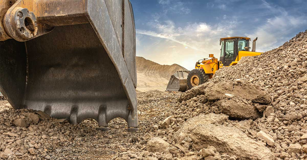 Close-up of a large excavator bucket in the foreground and a yellow front loader in the background, surrounded by mounds of dirt at an active construction site under a partly cloudy sky.