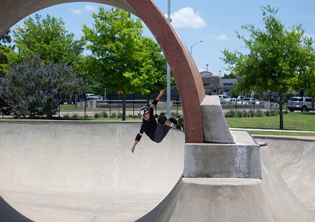 A skateboarder wearing protective gear rides along the curved wall of a concrete skate bowl beneath a large circular metal feature at the North Houston Skate Park.