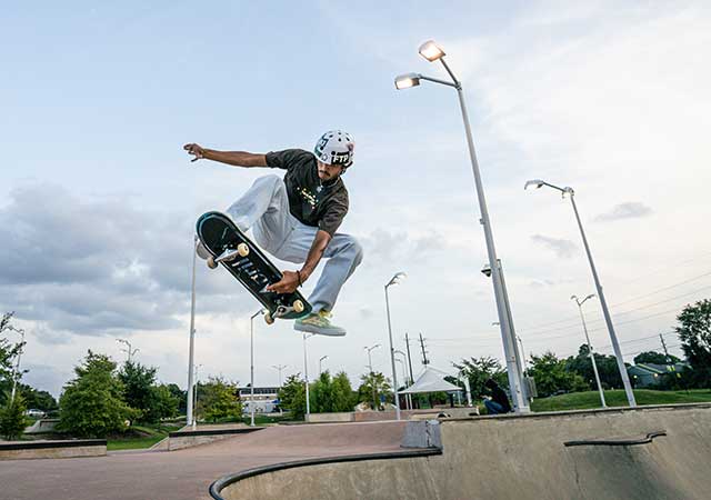 A skateboarder wearing a helmet performs a high jump above a concrete skate bowl at the North Houston Skate Park, captured midair against a cloudy sky and light poles.