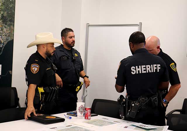Four uniformed officers from various law enforcement agencies stand in discussion near a conference table covered with meeting materials.
