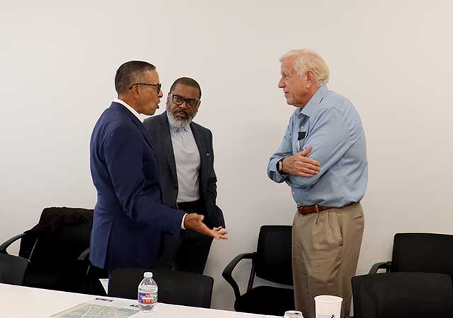 Three men engaged in conversation during a break at the Public Safety Committee Meeting, standing beside an empty row of chairs and a conference table.