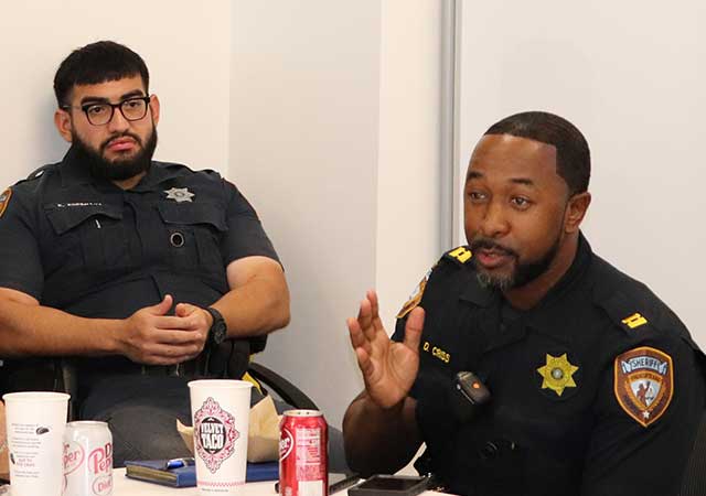 Two uniformed sheriff's deputies speak during the meeting, seated at a table with drinks and lunch containers.