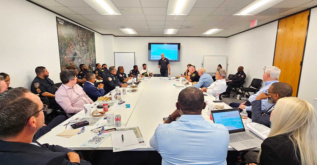 A presenter addresses a full room of public safety officers and community members seated around a large conference table beneath a wall-mounted screen.