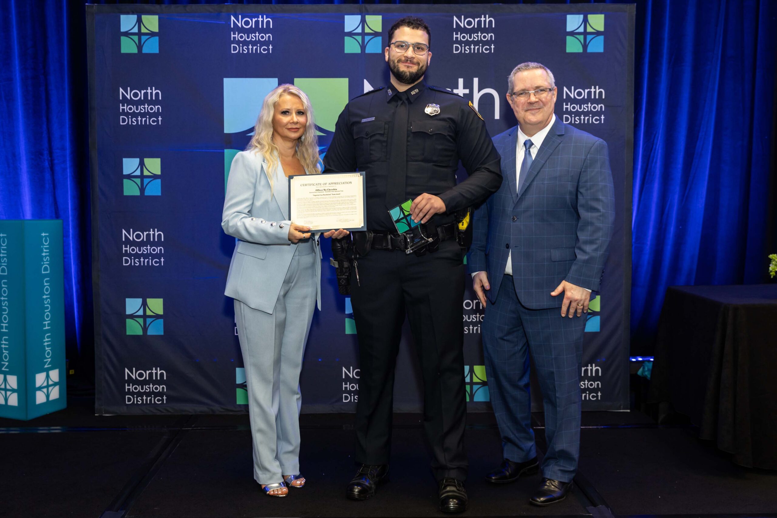 Public Safety Awards Luncheon 2025 Three people stand together smiling. A woman in a blue suit holds a certificate with a police officer in uniform, while a man in a blue plaid suit stands to the side.
