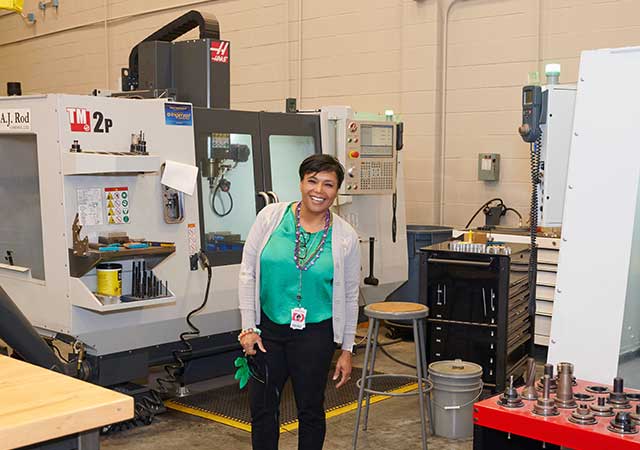 Wanda Ardoin Bailey 2 Wanda smiles in front of industrial CNC machinery in a classroom, wearing a green blouse and ID badge.