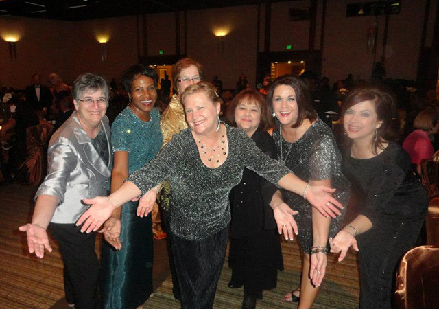 Seven women dressed in formal evening attire pose together with open arms and big smiles during the 2012 Woman of Excellence Gala.