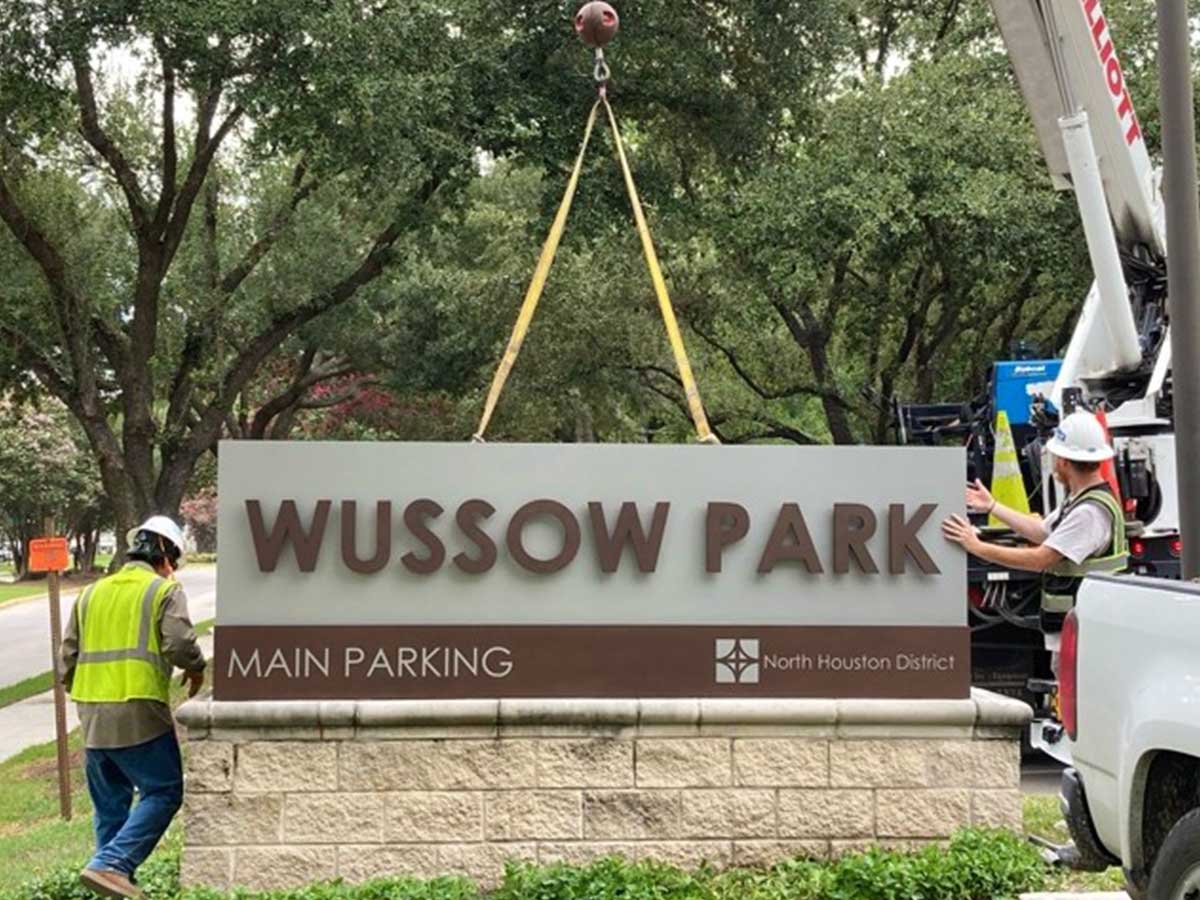 Construction workers install the new Thomas R. Wussow Park sign marking the main parking entrance. The sign displays the North Houston District logo and is surrounded by mature trees and park landscaping.