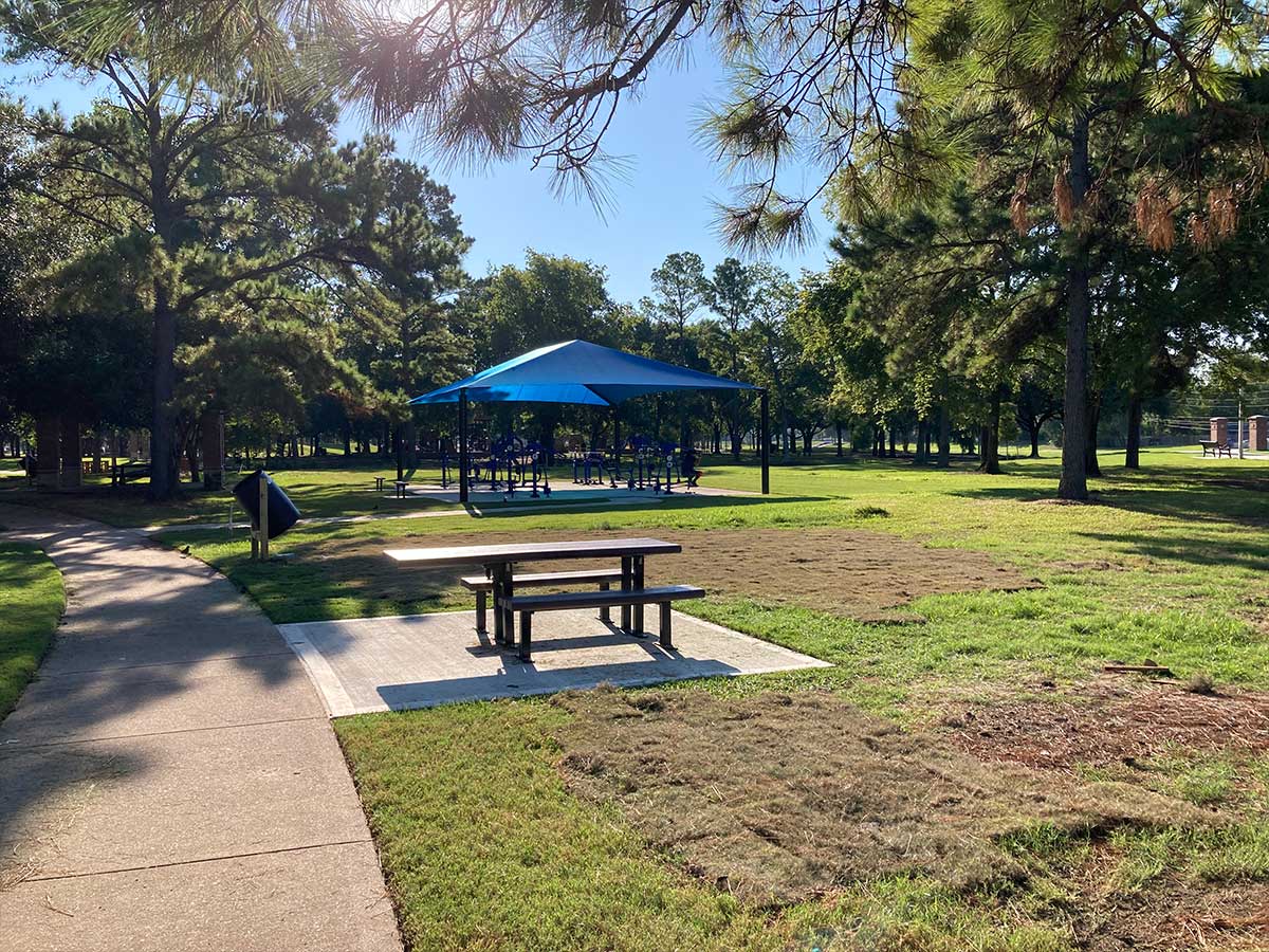 New picnic table and walking path at Thomas R. Wussow Park with a shaded blue fitness canopy in the background surrounded by tall pine trees and fresh landscaping.