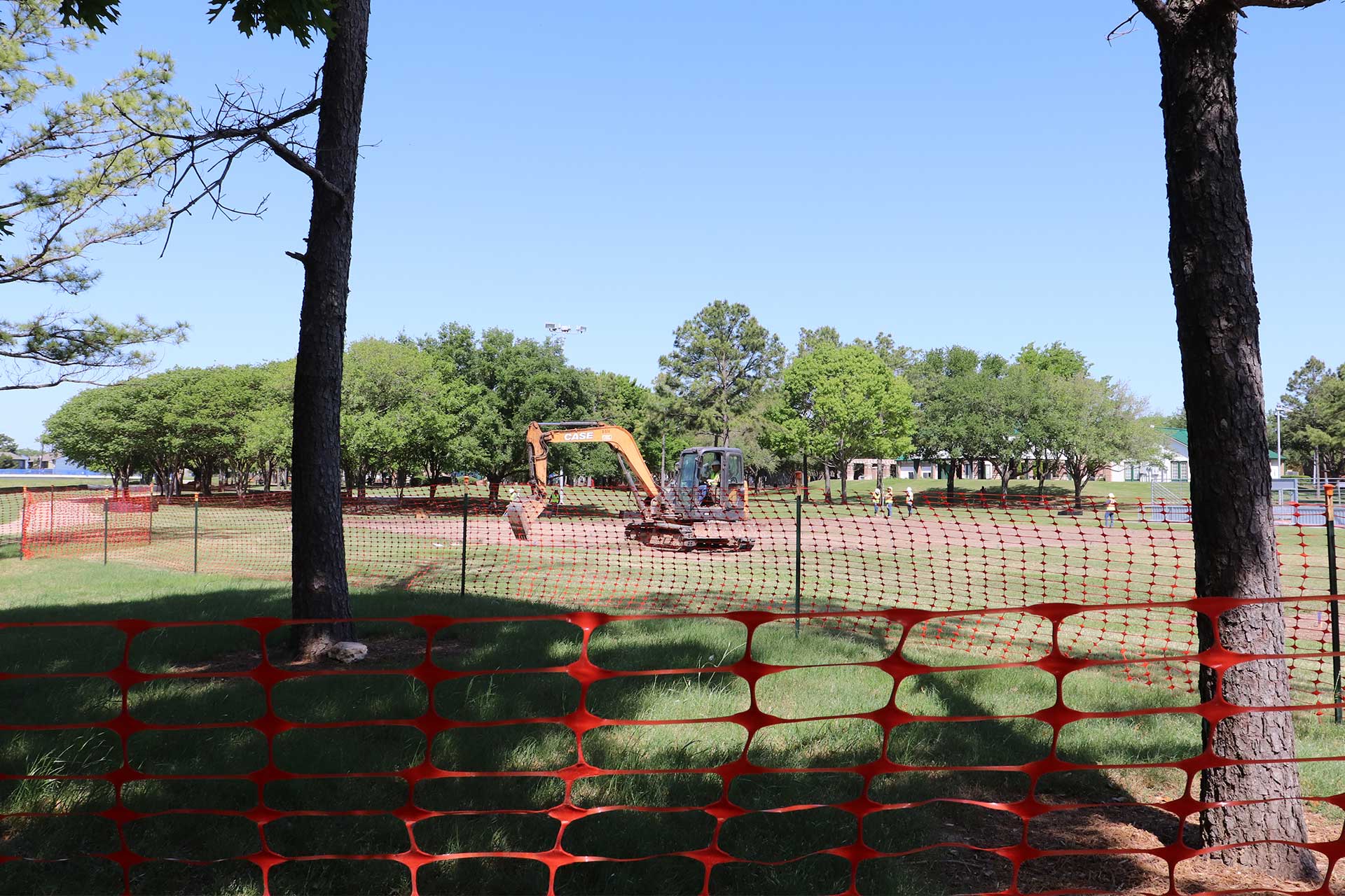 An orange safety fence surrounds a construction zone at Wussow Park, where a CASE excavator and workers are preparing a dirt-covered area for renovation. Trees and grassy areas frame the scene under a clear blue sky.